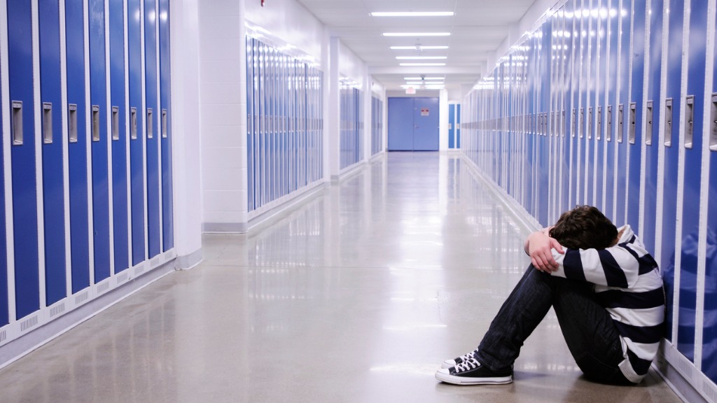 Fotografía de un joven sólo sentado en el suelo en medio de un pasillo de un centro escolar