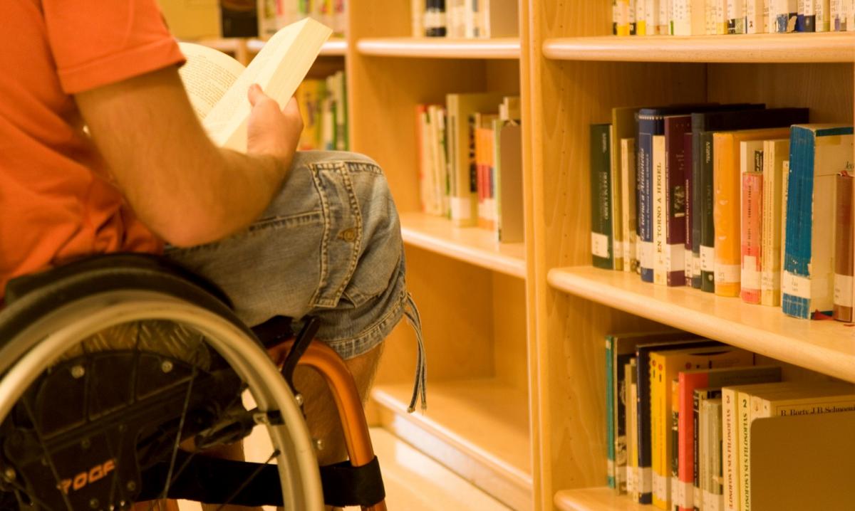 Una alumna universitaria en silla de ruedas consulta un libro en la biblioteca