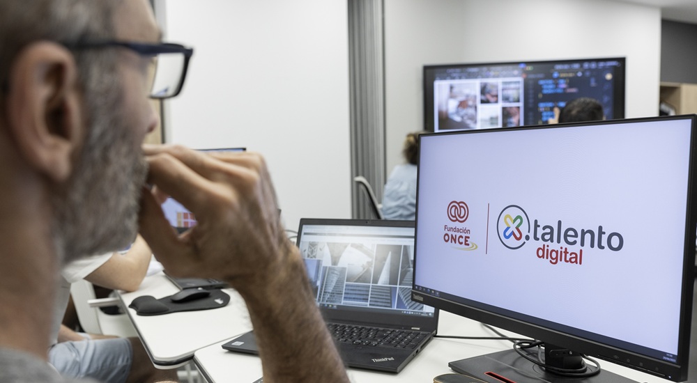 Un hombre en su puesto de trabajo junto a su ordenador portátil y frente a una pantalla en la que aparece el logotipo de Por Talento Digital