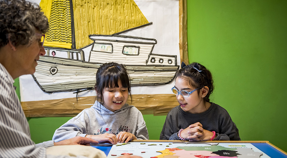 Dos alumnas con discapacidad visual en el aula junto a su profesora con un mapa de España sobre la mesa
